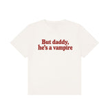 An off-white short-sleeve t-shirt with the phrase 'But daddy, he's a vampire' printed in red serif text across the chest, displayed on a solid white background.