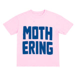 Pink t-shirt with blue text 'MOTHERING' on a white background