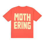 A coral red short-sleeve t-shirt with the word 'MOTHER' printed in large cream-colored block letters on the chest, with 'ERING' printed below it in the same style, spelling out 'MOTHERING'.